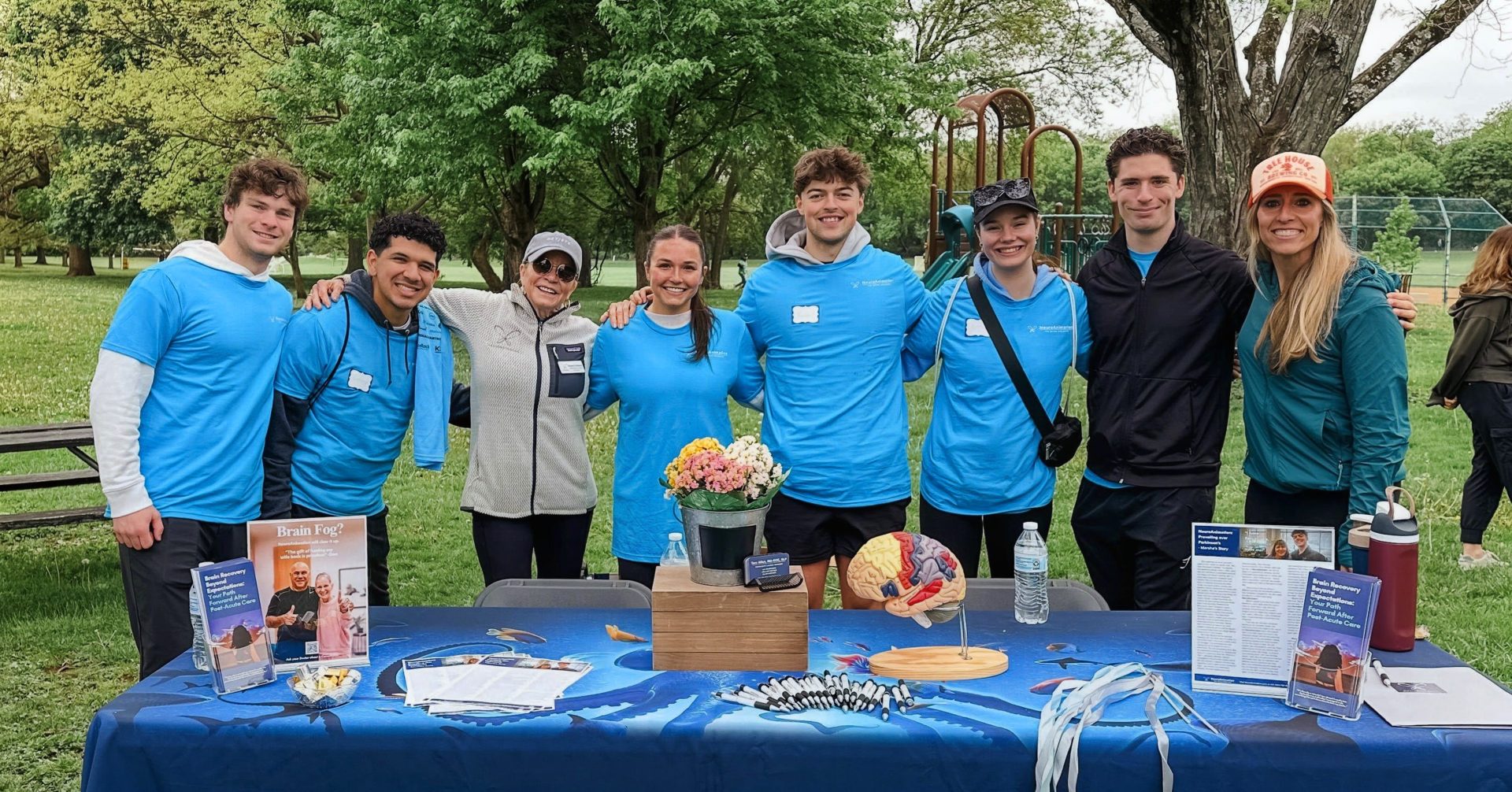 Eight people stand behind an information table at a community park event.